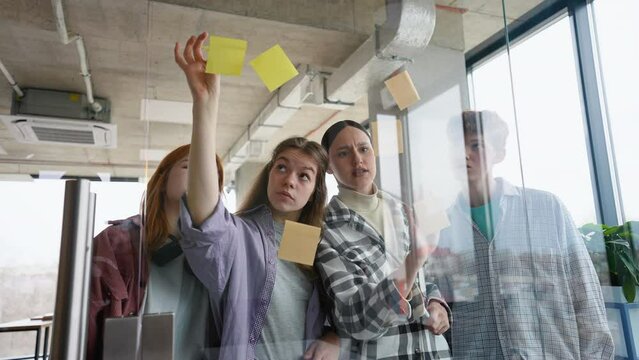 Group of students while working together during brainstorming and standing behind glass wall with sticky colorful papers