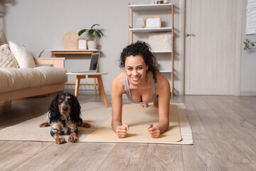 Sporty young African-American woman with cocker spaniel doing plank at home