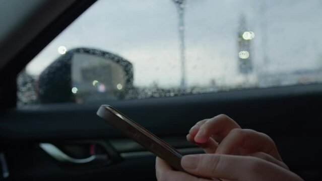 Hands of woman using smartphone in car, raindrops on window, city lights blurred in background
