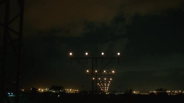 An airplane approaches for a nighttime landing, illuminated by runway lights against a dark sky, with approach lighting system visible in the foreground guiding the path