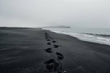 A lone path of footprints cuts through the stark, moody landscape of a black sand beach shrouded in mist