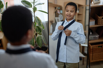Mirror reflection portrait of young Black boy tying tie and smiling while dressing up at home
