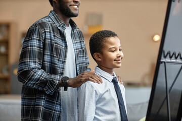 Side view portrait of young Black father and son looking in mirror together while dressing up at home