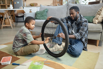 Side view portrait of Black young father and son fixing bicycle wheels together at home
