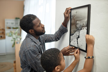 Side view portrait of African American father and son hanging picture on wall together after moving in new house - obrazy, fototapety, plakaty
