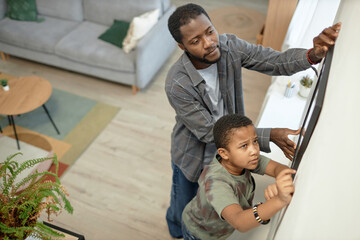 High angle view of African American father and son hanging picture on wall together decorating home