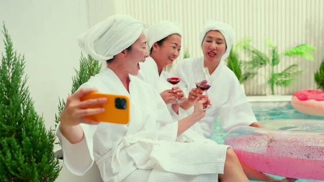 chilling sunday pool party asian adult friend woman in bathrope casual cloth hanging around sit near water pool hand hold glass wine enjoy selfie video calling old friend meeting laugh smile summer