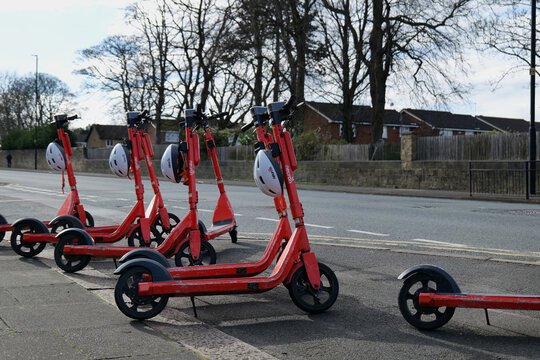 Red neuron rentable electric scooters lined up on a pavement in Newcastle. White safety helmets are hung on them