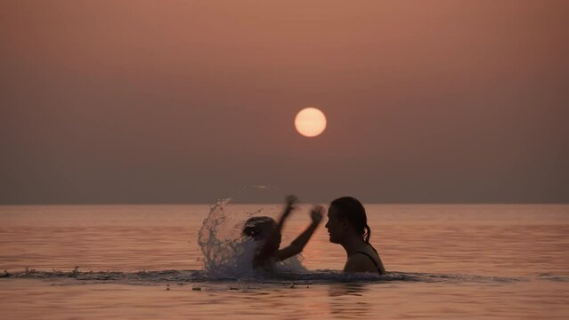 Mother in a swimsuit lifting and tossing her daughter into the air, with water splashing around them at sunset