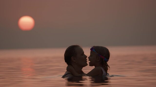 Close-up of a mother and daughter with their lips touching, wearing swimwear and goggles, in the sea with the setting sun in the background