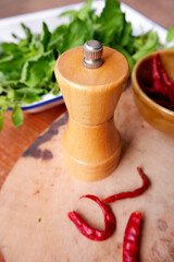Obraz na płótnie Canvas Pepper bottle with food preparation of holy basil and dried chili. The concept of equipment, cooking, and rustic kitchen. Close-up pepper grinder placed on wooden cutting board. Food vertical image.