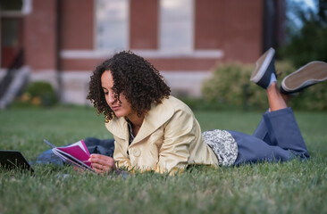 African-American female student studying while laying on lawn in front of college building in USA; notebook and laptop computer in front of her