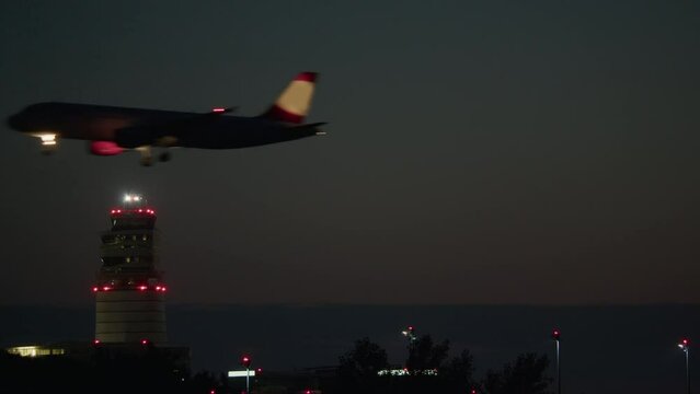 An airliner with illuminated landing lights flies low at night over a control tower with red warning lights