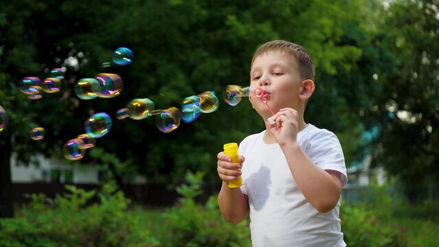 Little joyful boy in t-shirt blows soap bubbles playing in summer city park. Cheerful boy blows bubbles in park among trees on vacation. Schoolboy spends time blowing soap bubbles slow motion