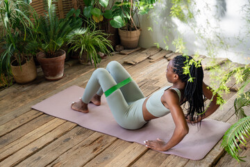 Active african american woman doing exercises with fitness elastic band sitting on sported yoga mat surround by indoor plants in sunny day. Athletic black young female home training for healthy body.