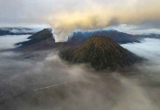 An ethereal aerial view of volcanic peaks emerging from a sea of mist at dawn, with smoke rising from an active crater