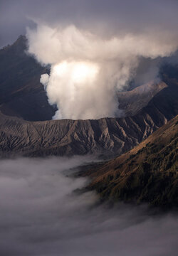 The morning light accentuates a volcanic eruption, its plume glowing above the mist-filled valleys, showcasing the raw beauty of nature