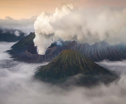 A breathtaking view captures the volcanic eruption's smoke rising above the mist-shrouded mountains at dawn, with soft light caressing the scene