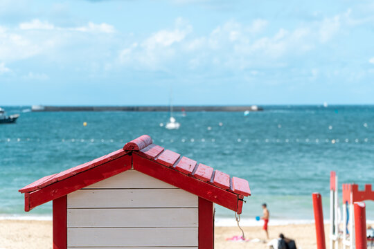 Vivid red beach hut stands out against the serene blue waters, with boats sailing in the background in Saint Jean de Luz, France