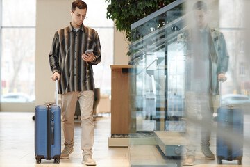 Full length portrait of modern young man with suitcase walking towards camera in hotel lobby and using smartphone with taxi app copy space - obrazy, fototapety, plakaty