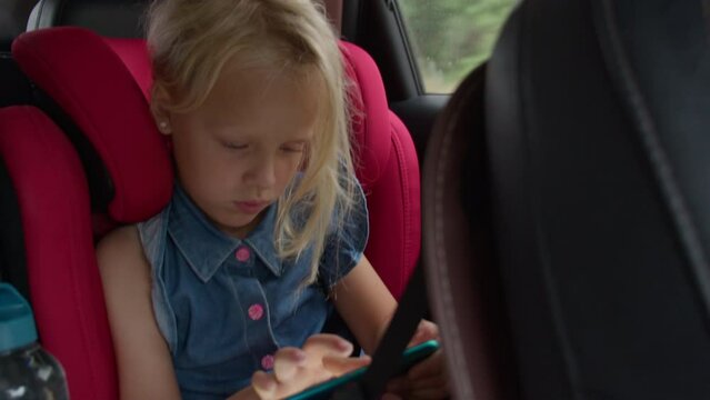A young girl is engrossed in a tablet while sitting in a bright red car seat, with the interior of the vehicle and a water bottle also in view