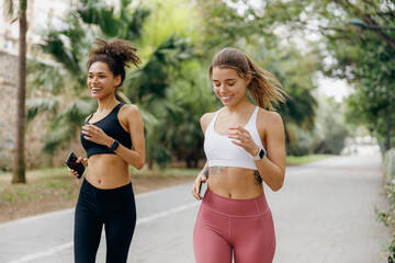 Two smiling women in sportswear are running on natural parkland background. Active lifestyle concept - obrazy, fototapety, plakaty