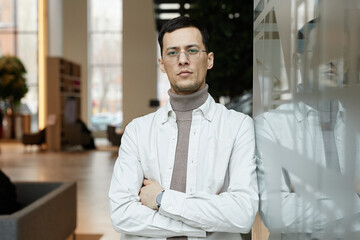 Minimal waist up portrait of confident Asian man wearing glasses leaning on glass wall in office...