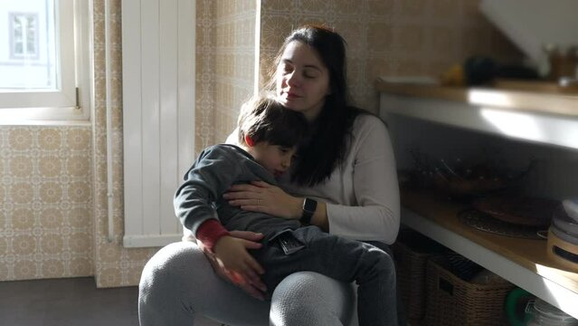 Mother resting while holding 5 year old son at kitchen, authentic family lifestyle scene of tired mom with eyes closed and kissing child in forehead in affectionate moment