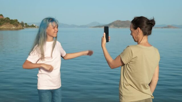 The young blogger is helped by his mother. A teenage girl with blue hair is dancing on the sea beach, mom is filming a dance on a smartphone.