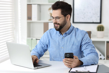 Handsome young man using smartphone while working with laptop at white table in office