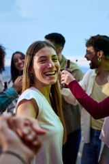 Obraz premium Vertical. POV young smiling Caucasian woman holding hands boyfriend at party. Attractive girl looking cheerful on camera friends in background outdoors. People gathered on rooftop celebrating birthday