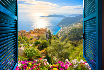 View through an open window with blue shutters and flowers of the hilltop medieval old city of Eze, France, with the Mediterranean coastline and see below.