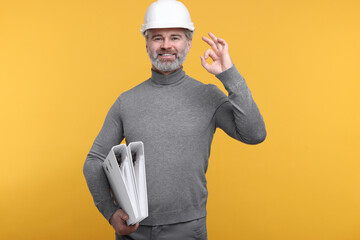 Architect in hard hat holding folders and showing ok gesture on orange background