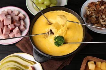 Dipping different products into fondue pot with melted cheese on black wooden table, above view