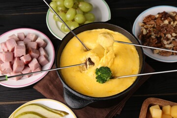 Dipping different products into fondue pot with melted cheese on black wooden table, above view