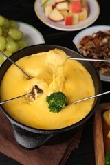 Dipping different products into fondue pot with melted cheese on table, above view