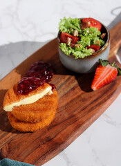 A portion of fresh made fried camembert cheese with cranberry sauce on top and a side of fresh salad on wooden plate