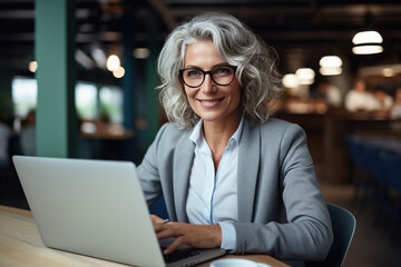 A poised senior businesswoman with sophisticated silver curls confidently uses her laptop in a bright, modern office environment, her professional attire speaking to her experience - obrazy, fototapety, plakaty