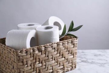 Toilet paper rolls and green leaves in wicker basket on white marble table, space for text