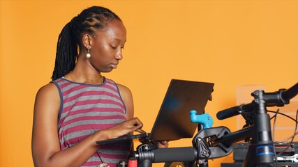 Cheerful mechanic doing bike servicing in studio background atelier shop, looking on laptop maintenance list. Specialist checking online for replacement bicycle parts using notebook, camera A