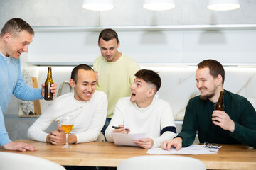 Group of cheerful young adult men gathered at cozy home kitchen, enthusiastically toasting with beer to celebrate new job offer of friend receiving paper notice