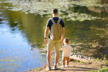 A young father walking with his toddler son along the shore of a pond or river on summer day. A baby boy is learning to walk.