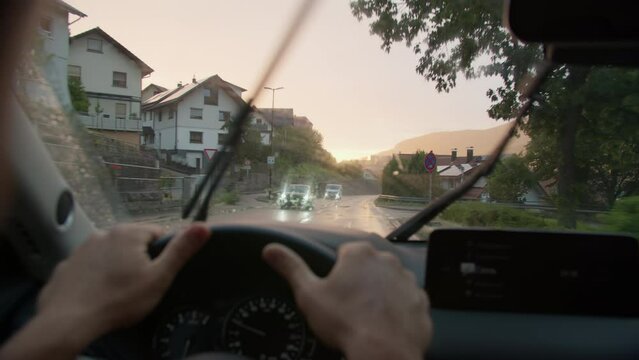 Driver's view from inside a car with hands on the steering wheel, windshield wipers active, on a wet road reflecting the sunset in a residential neighborhood