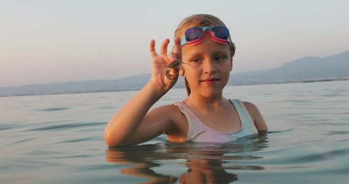 A girl in a swimsuit and swimming goggles stands in the sea at sunset, holding a small crab with her fingertips