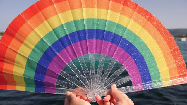 A hand-held rainbow fan glitters under the sunlight, with a backdrop of blue water, symbolizing LGBT pride and diversity