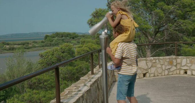 A boy in a striped shirt lifts a girl in a yellow dress, onto his shoulders so that she can look through a stationary open telescope at a pond with distant hills