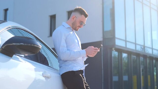 A slender business man in sunglasses is standing looking at the phone next to the expensive car.