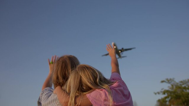 Mother and daughter stand with their backs to the camera and wave to a passing plane