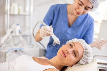 Obraz na płótnie Canvas Young woman cosmetologist performs facial moisturizing procedure with machine to young female client