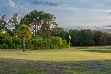 A yellow flag stands motionless attached to a pole in the center of a golf green. The background...